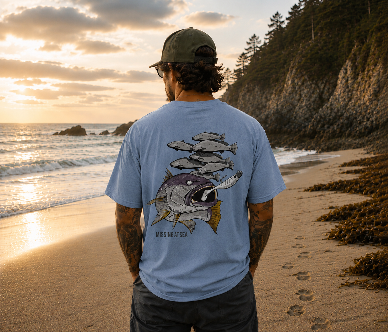 Man standing on a beach wearing a light blue t-shirt with a fish graphic and text, looking at the sunset.