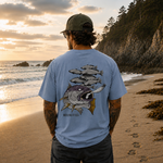 Man standing on a beach wearing a light blue t-shirt with a fish graphic and text, looking at the sunset.