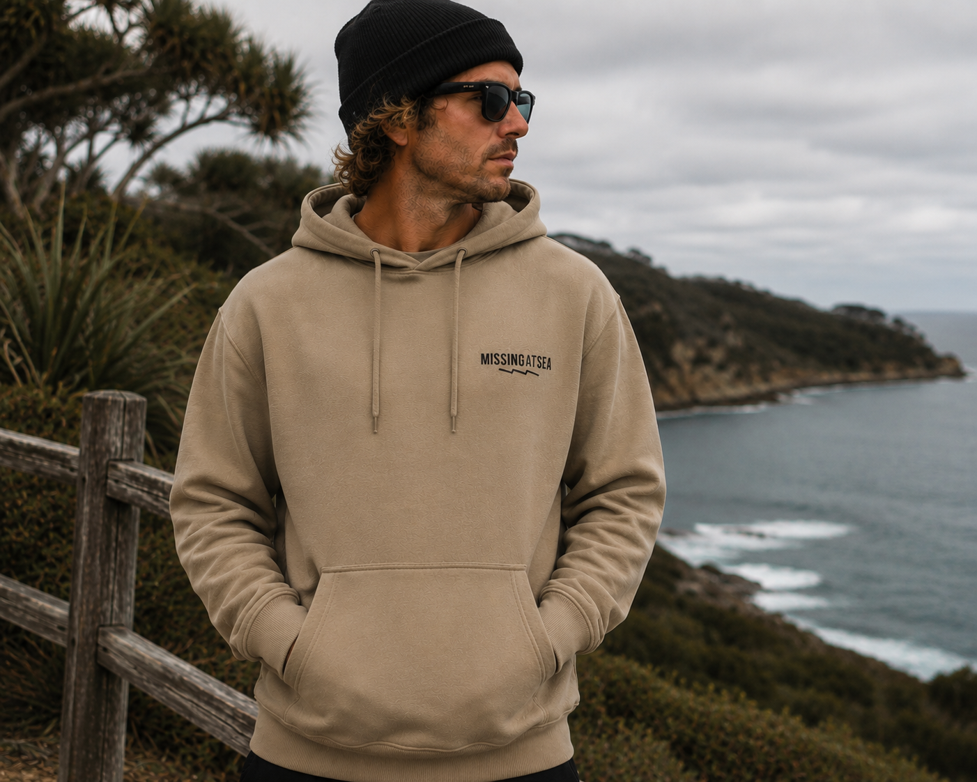 Man wearing a beige hoodie with a logo, standing on a coastal landscape.
