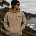 Man wearing a beige hoodie with a logo, standing on a coastal landscape.