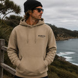Man wearing a beige hoodie with a logo, standing on a coastal landscape.