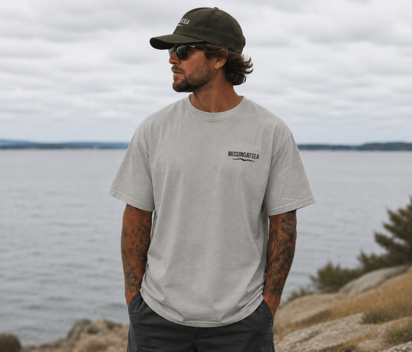 Man wearing a gray t-shirt with text and a cap, standing by a body of water.