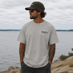 Man wearing a gray t-shirt with text and a cap, standing by a body of water.