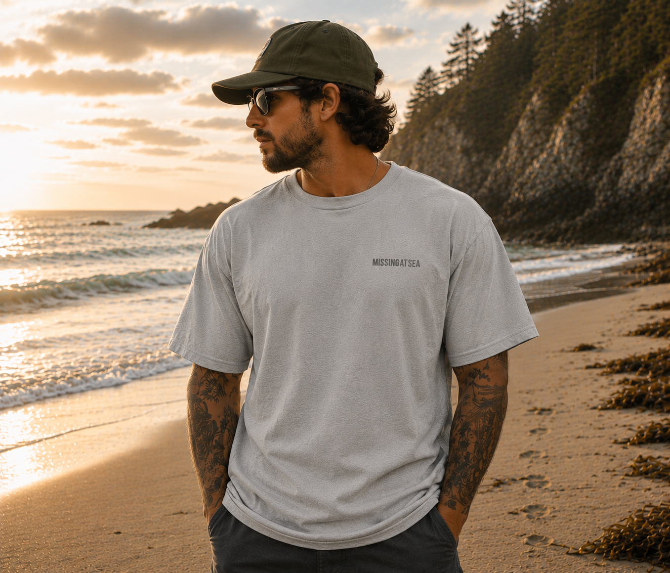 Man standing on a beach at sunset wearing a gray t-shirt and cap.