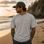 Man standing on a beach at sunset wearing a gray t-shirt and cap.