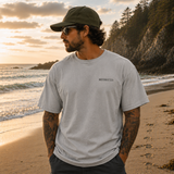 Man standing on a beach at sunset wearing a gray t-shirt and cap.