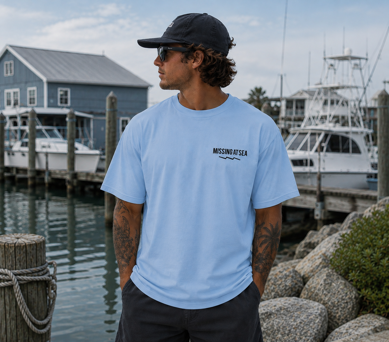 Man wearing a light blue t-shirt with 'Missing at Sea' logo by a marina.