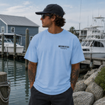 Man wearing a light blue t-shirt with 'Missing at Sea' logo by a marina.