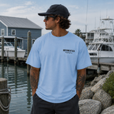 Man wearing a light blue t-shirt with 'Missing at Sea' logo by a marina.