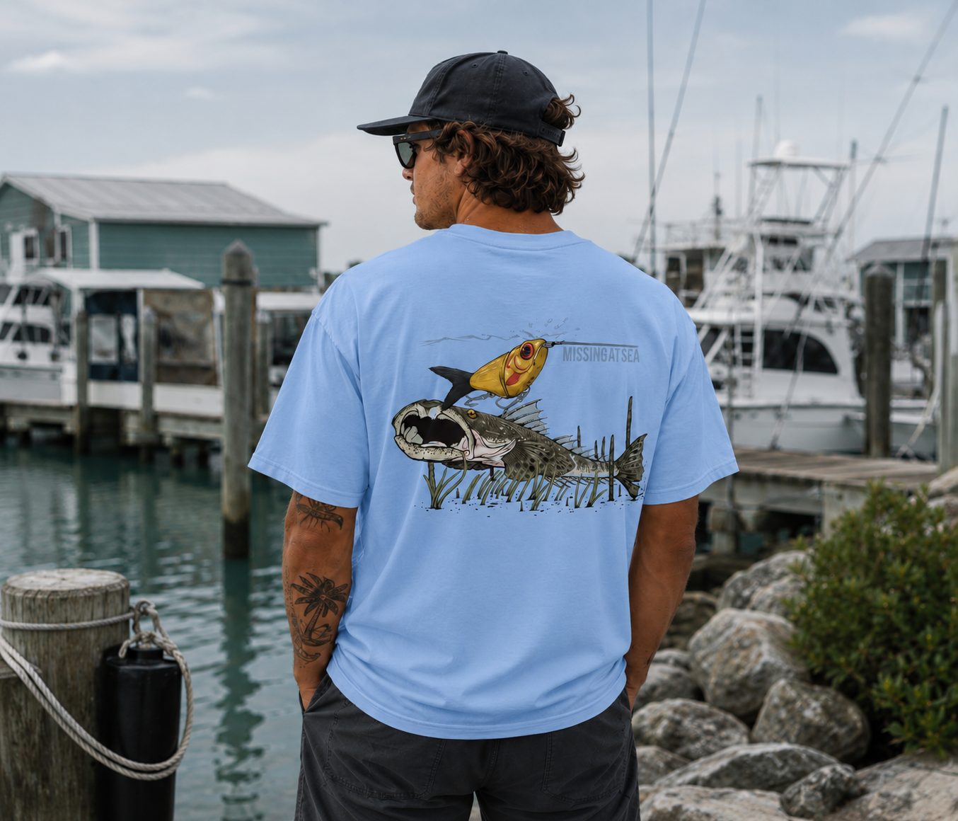 Man wearing a light blue t-shirt with fish graphics by a marina.